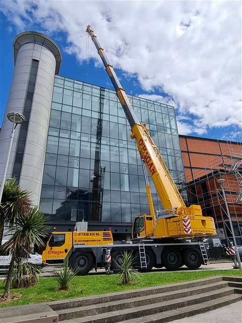 Side view of yellow five axle all terrain crane, boom up in front of a big glass building
